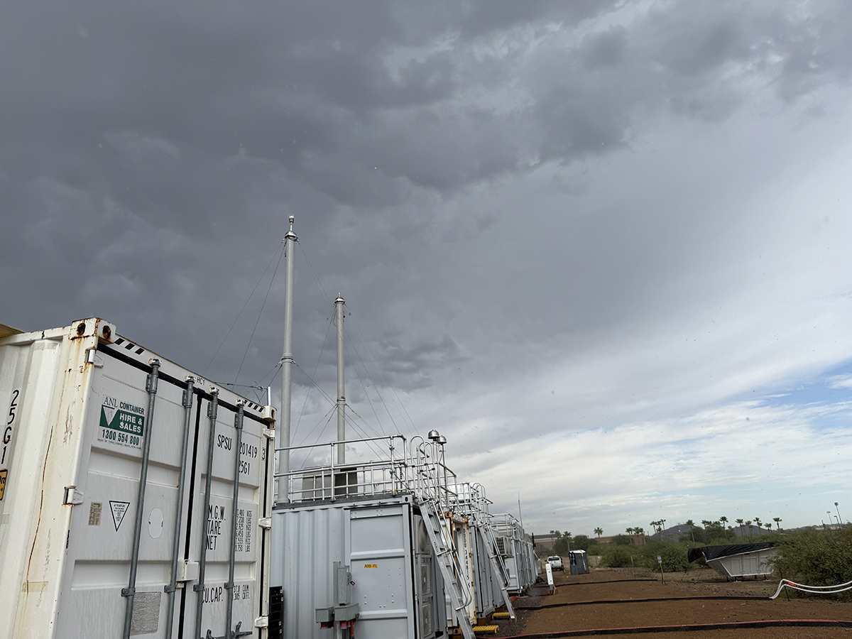 Dark clouds sit over large containers and atmospheric instrumentation, including a pair of aerosol sampling stacks, with shrubs, palm trees, and a modern academic building nearby.
