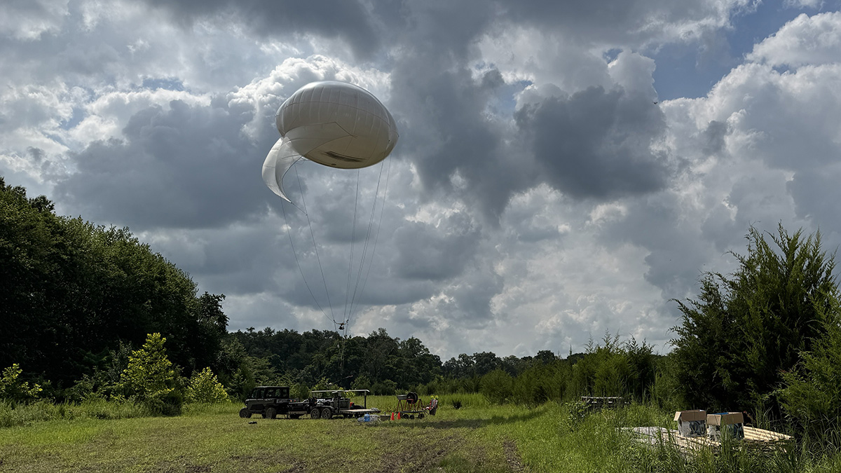 A white balloon attached to a tether is seen amid a cloud-filled sky. The balloon platform and truck are parked in an open field surrounded by trees and bushes.