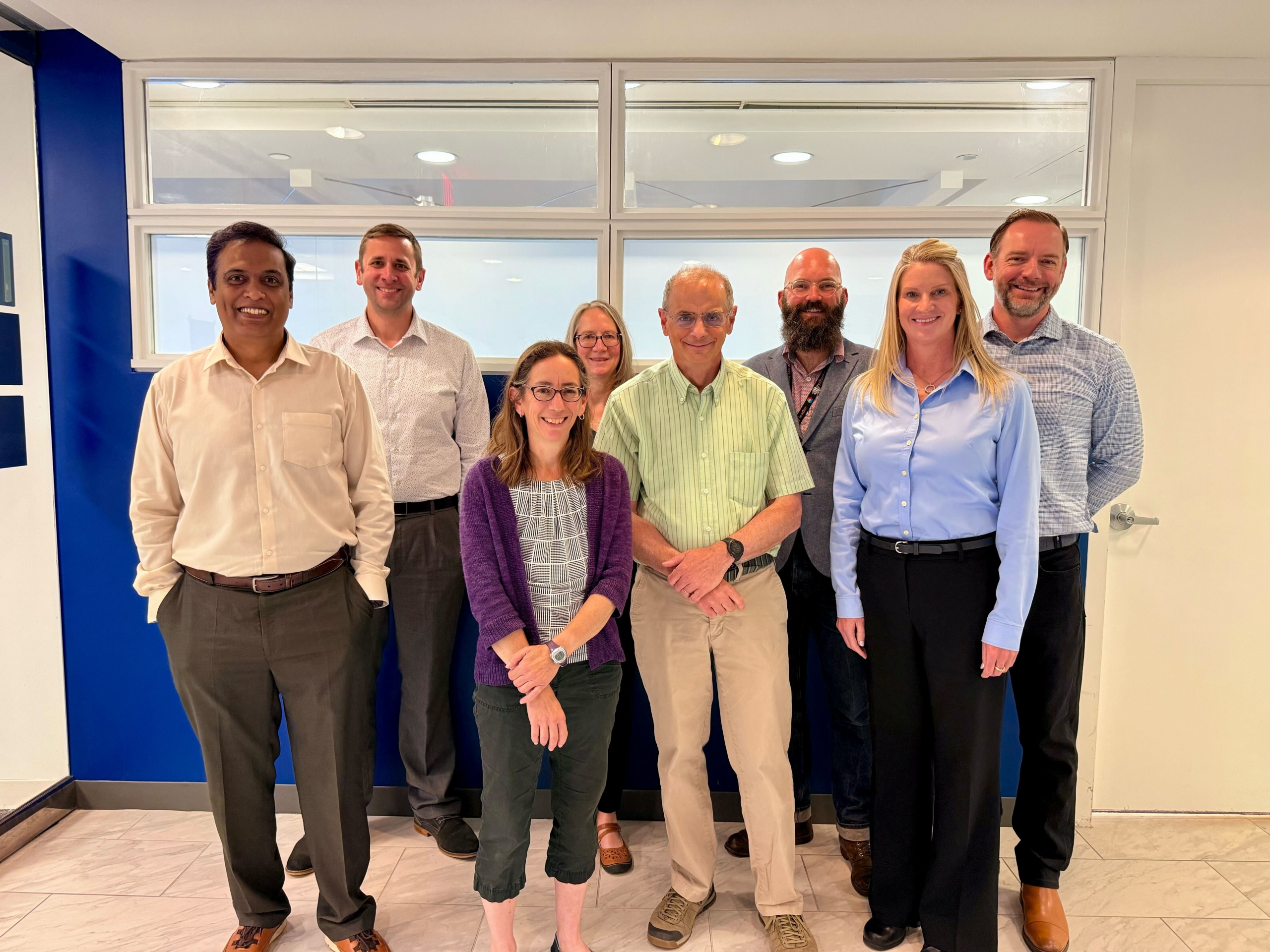 A group of people gathered for a business meeting stand and smile for a photo in front of a window and blue wall.
