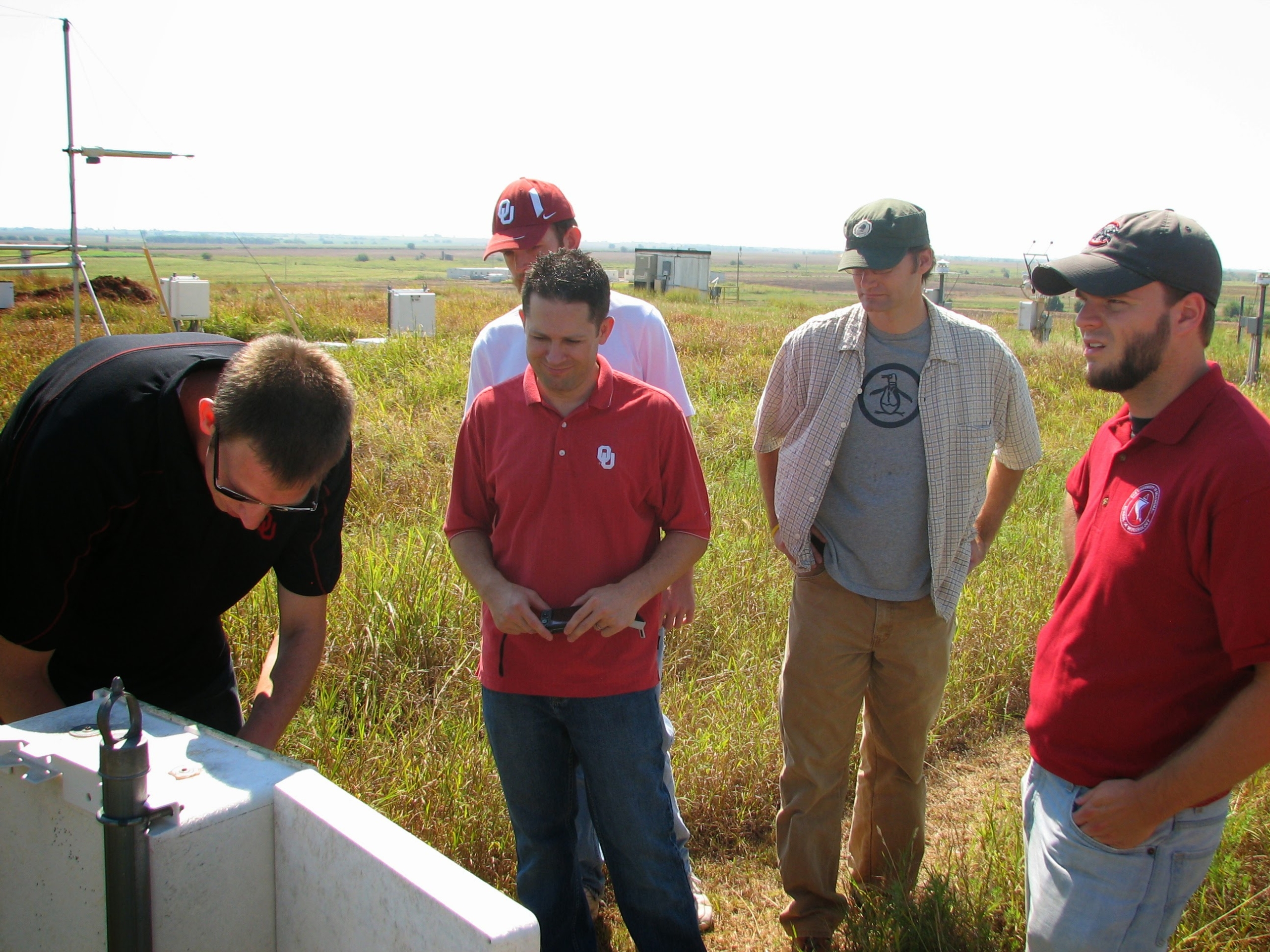 Four people look on in a grassy field while another person leaning down works on an instrument.