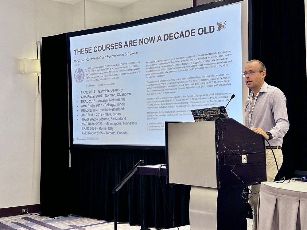 A person stands at a podium with a laptop open and a large screen to their side that shows a schedule for the past 10 years of ERAD/AMS open source radar software short courses.