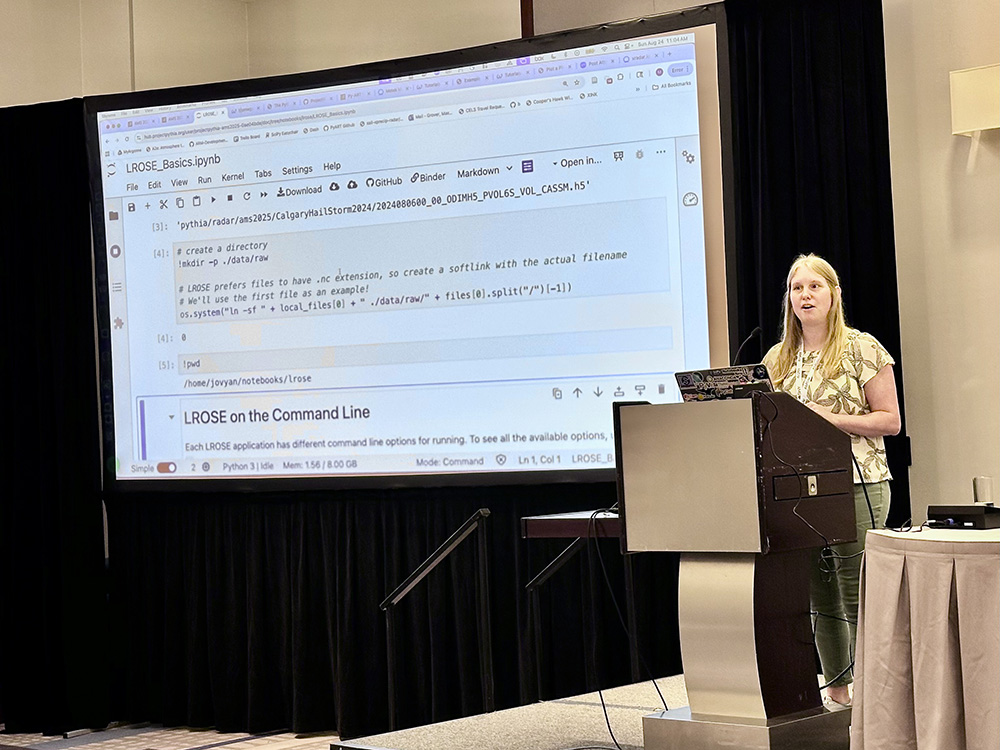 A person stands at a podium with their laptop open and a large screen to their side showing LROSE code.