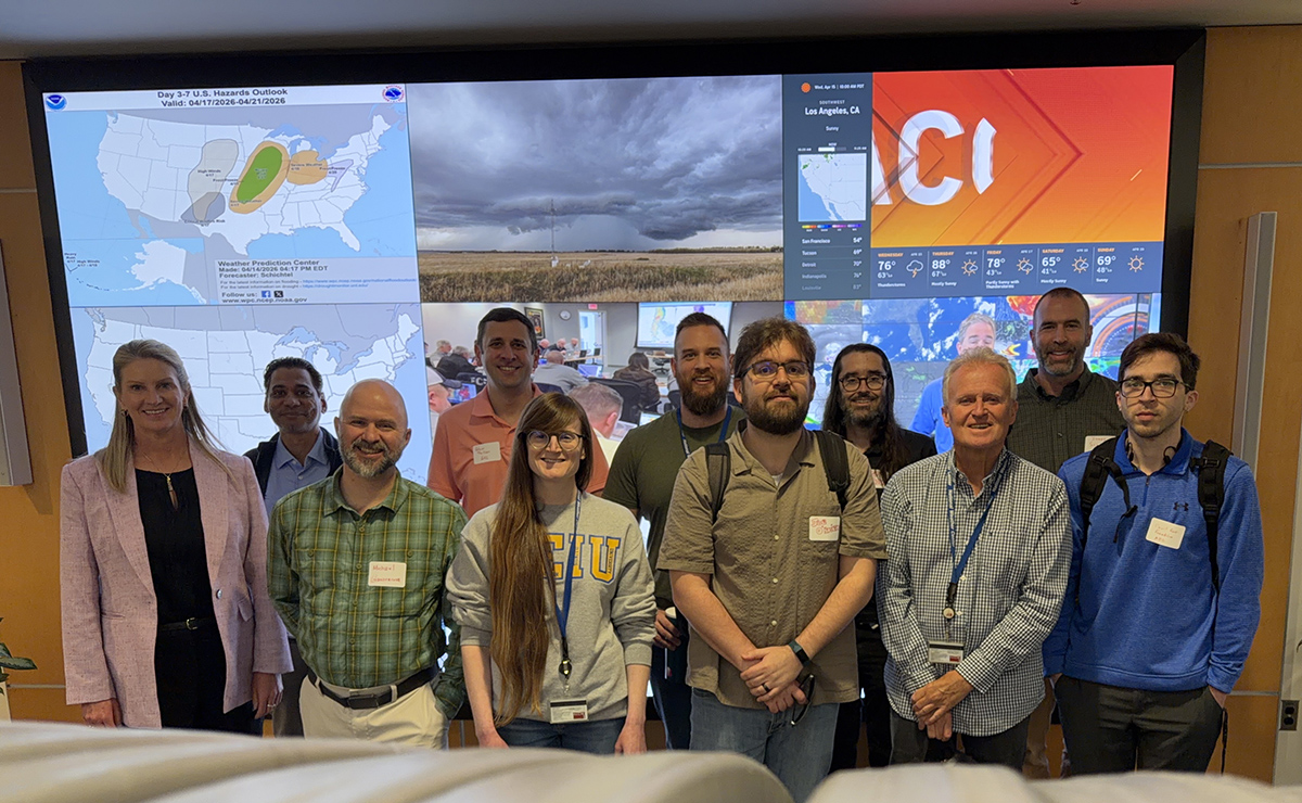 Members of ARM's new data quality operations organizational chart pose for a picture in front of a large screen with six different images showing. Fully visible images include a U.S. hazards outlook map, storm clouds, and icons illustrating weather in different cities.