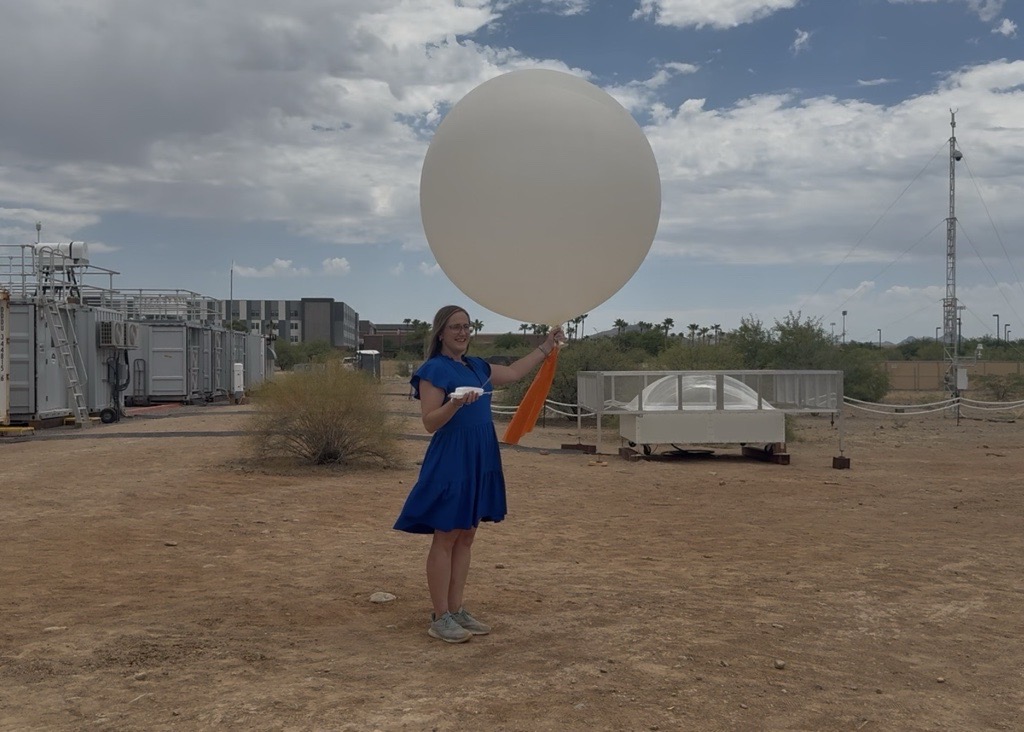 A person wearing a blue dress and glasses holds a radiosonde in her right hand and a weather balloon in her left. The person is standing on dirt and surrounded by atmospheric instrumentation and large containers under a partly cloudy sky. A modern academic building and desert vegetation are seen behind the person.