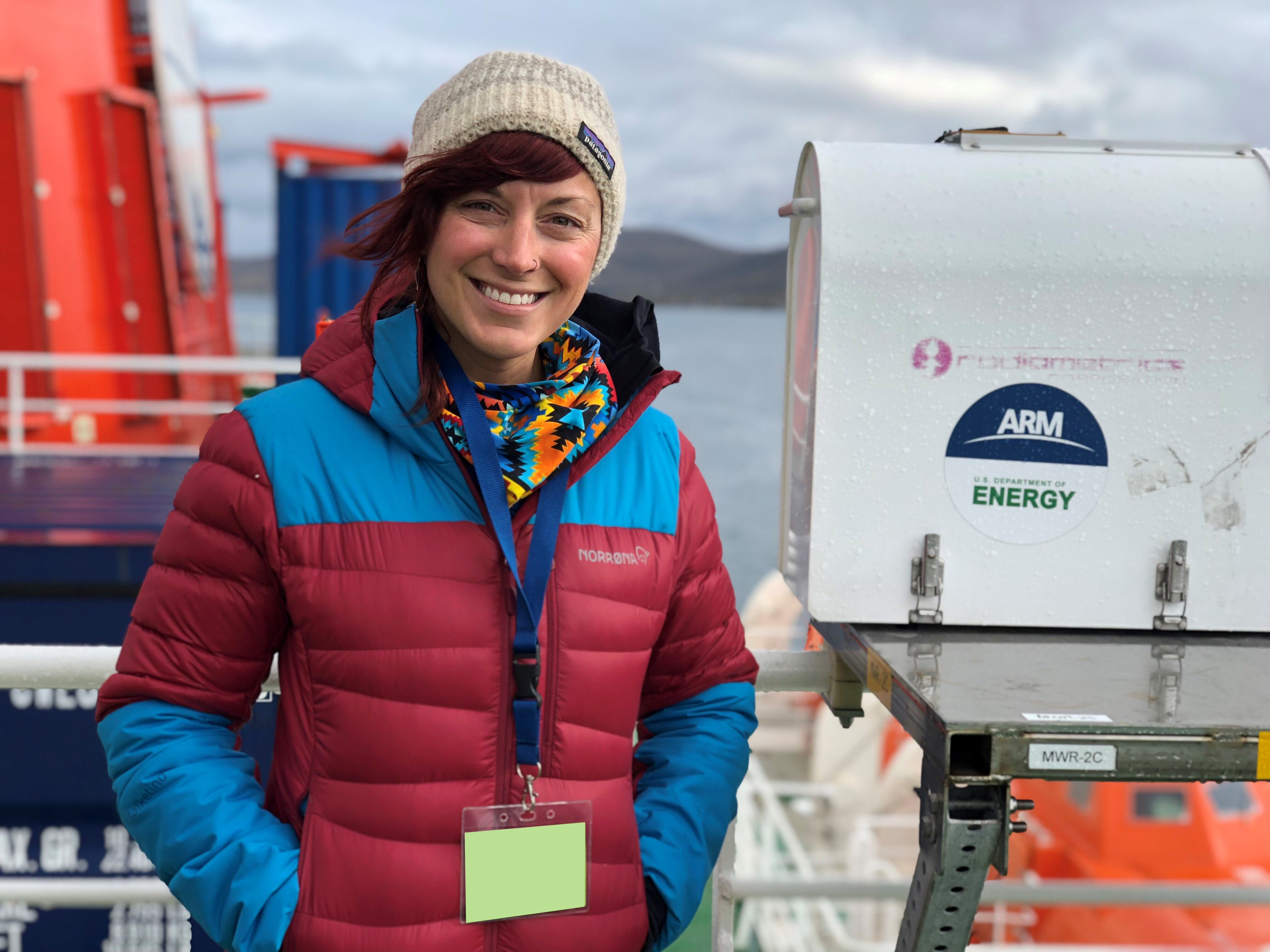 Jessie Creamean smiles while standing next to a 2-channel microwave radiometer with an ARM/U.S. Department of Energy sticker on its side.