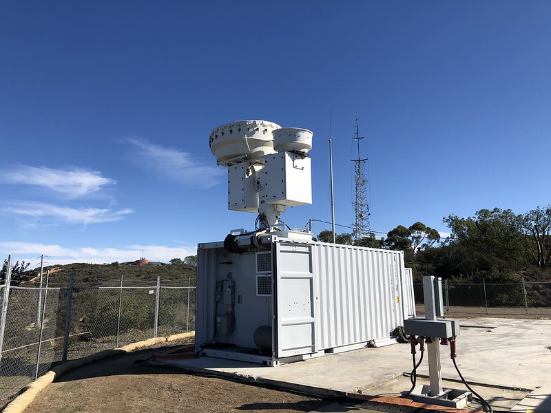 On a mostly sunny day with some clouds streaking across the sky, an instrument container with radars on top is sited on a concrete platform. Chain link fencing surrounds the instruments, with bushes and trees on the other side of the fence.