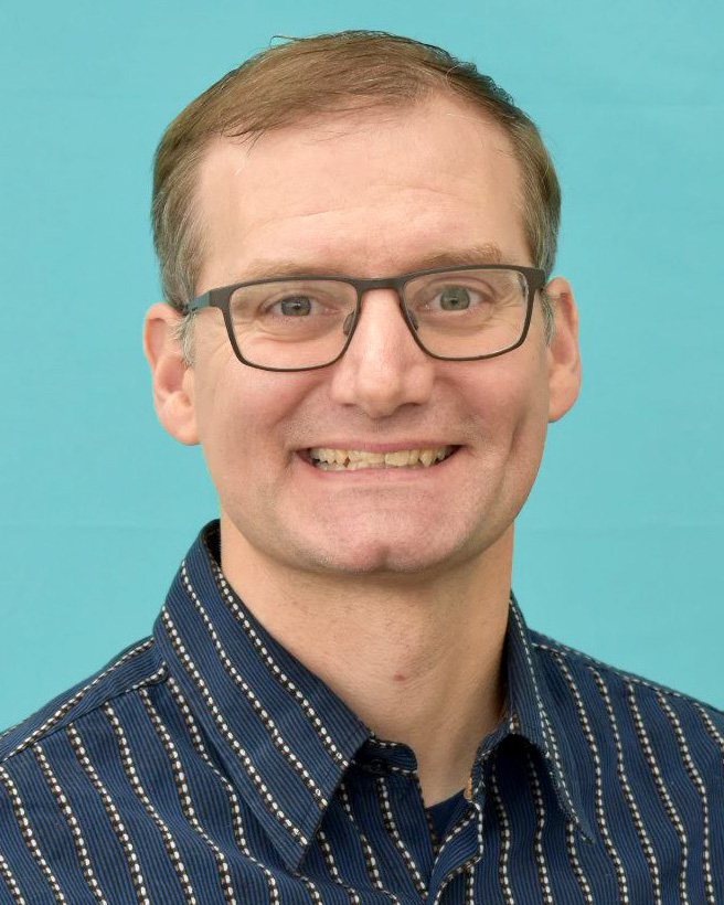 Portrait of Ken Kehoe wearing a striped shirt in front of an aqua background