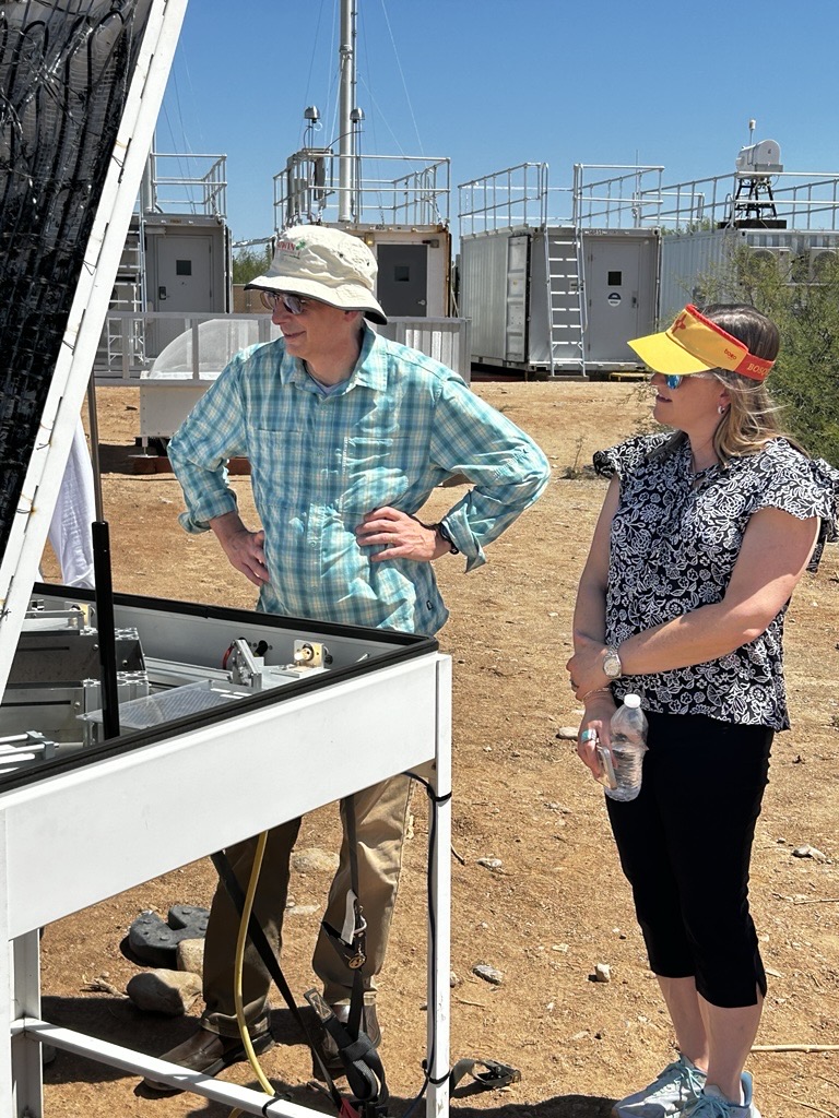 Two people stand in front of an open video disdrometer. One of the people has their hands on their hips, while the other is standing with one hand holding their arm. They are both smiling and appear to be listening to someone standing next to them out of the frame. Atmospheric instrumentation and containers are seen behind them.