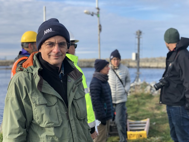 Jim Mather wears an ARM beanie and green jacket and looks at the camera on a cold, cloudy day with others in the background wearing beanies and jackets. A person behind Jim is wearing a safety vest and yellow protective helmet.