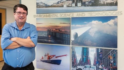 Photo of Mike Jensen in blue shirt with crossed arms standing in front of a photo gallery wall with the text "Environmental Science and Technologies"