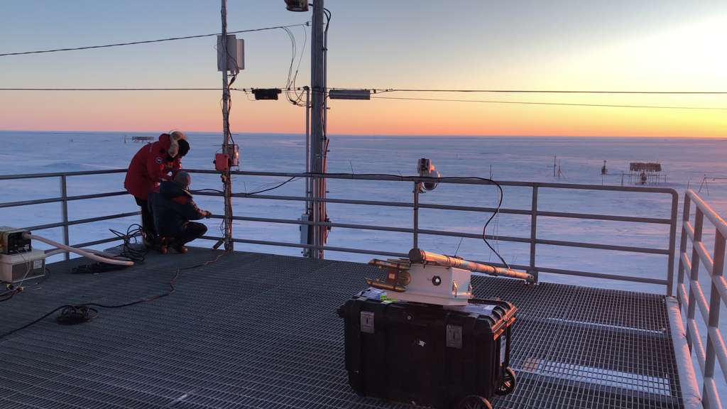 Two people work together in a corner of an instrument deck with the sun illuminating the sky from the right side of the photo. Over the instrument deck railing, instruments are seen out on the snow.