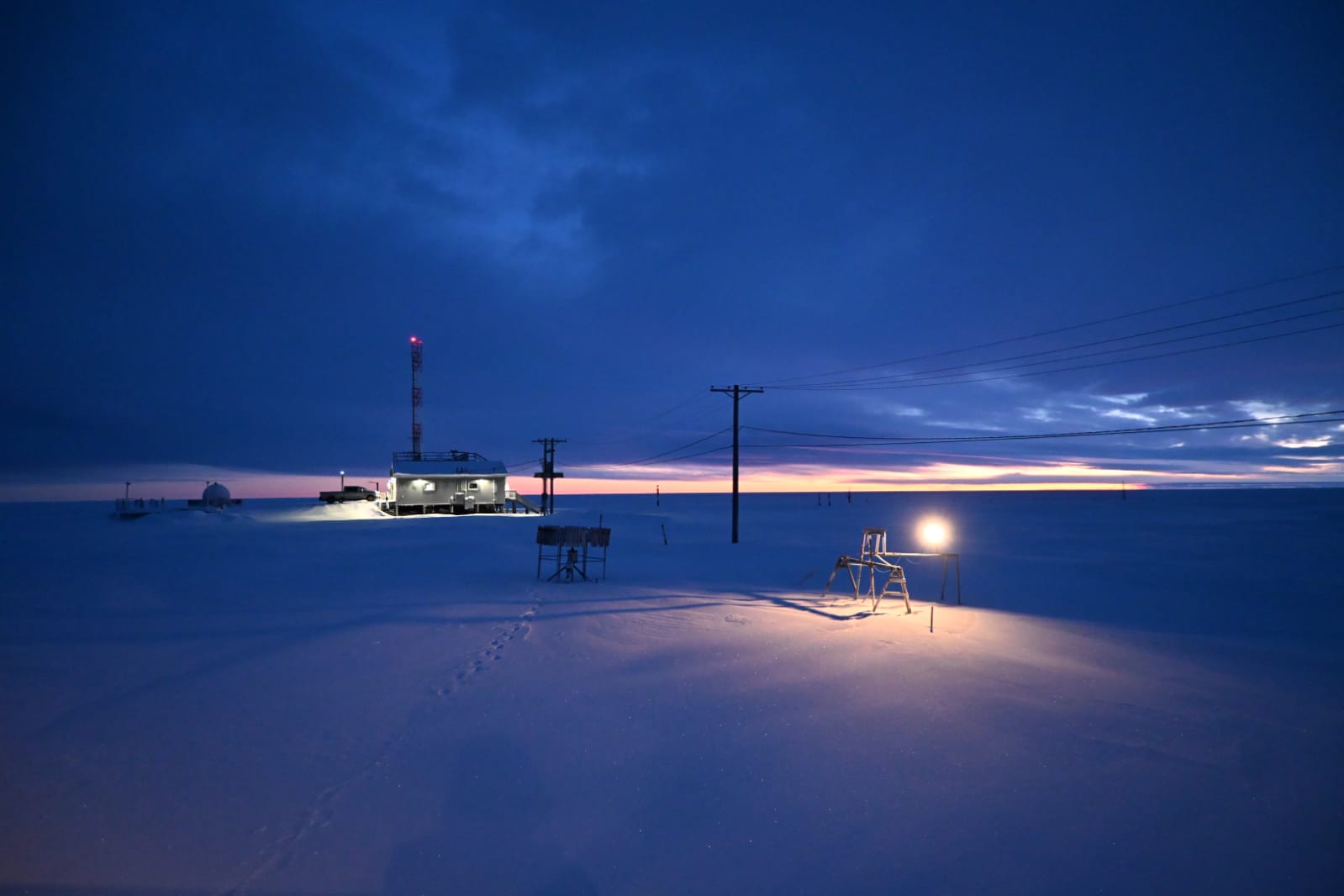 A building, a rain gauge, and a laser precipitation probe (with a light source and camera) are seen with the sun barely peeking over the horizon and a blanket of snow on the ground. A trail of footprints is visible in the snow.