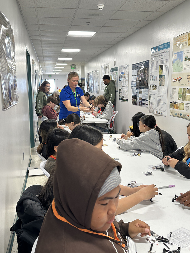 Helena Buurman (blue shirt) from the Office of the Vice Chancellor for Research, Alaska Satellite Facility at the University of Alaska Fairbanks, teaches children in Utqiaġvik, Alaska, how to assemble small uncrewed aircraft at the Barrow Arctic Research Center (BARC) Science and Culture Fair in August 2024. Photo is courtesy of Valerie Sparks, Sandia National Laboratories.