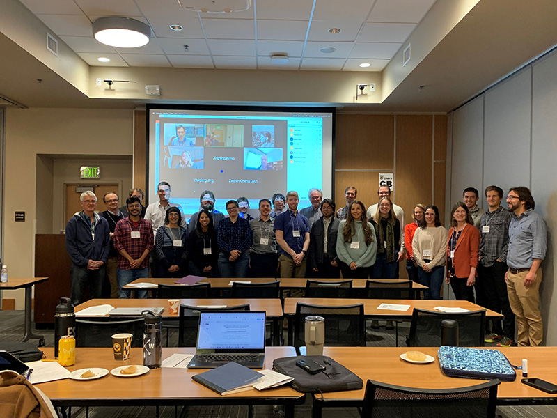 A group of people pose in front of a screen featuring virtual meeting attendees.