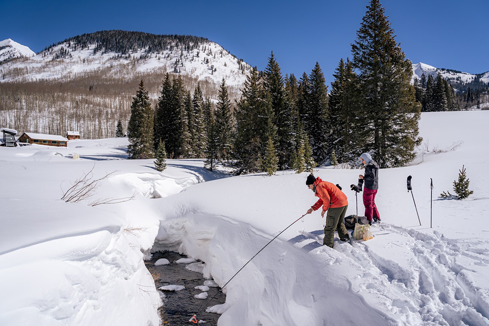 One person sticks a probe into an icy stream while another person overlooks the process. Snow blankets the ground, mountains, and roofs of nearby small wooden cabins. Trees stick out relatively far above the snow.