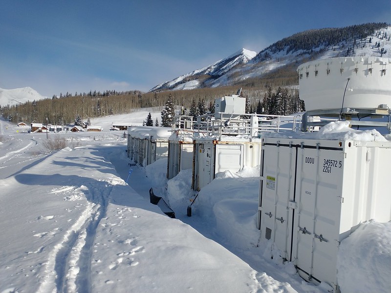 Snow is piled up in between large instrument containers, and ski tracks cut through snow in front of the containers. Trees, buildings, and a snow-covered mountain are visible in the distance on a mostly sunny day.
