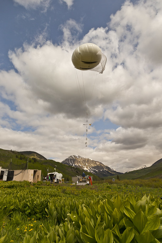 A large balloon with instruments on a tether hovers in the sky over a leafy field with some yellow flowers peeking through in the corner. A mountain with sparse snow cover is seen in the distance with clouds overhead.