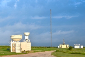 Atmospheric instruments and radars are pictured on a green field with a blue sky and white clouds
