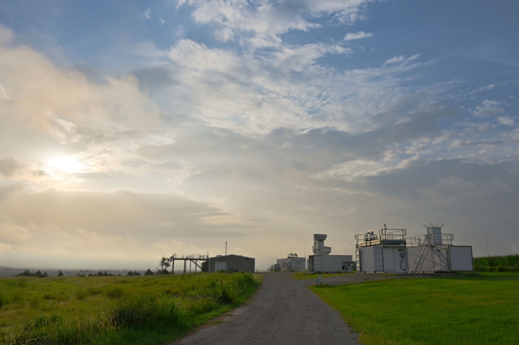 A blue sky with clouds and sunlight are pictured above atmospheric instruments and a gravel road