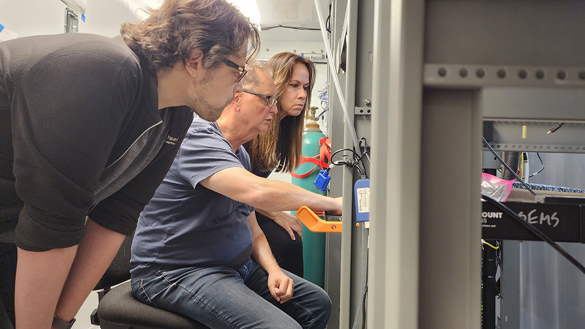 Two people bend down to look at aerosol instrumentation with a person sitting in the middle.
