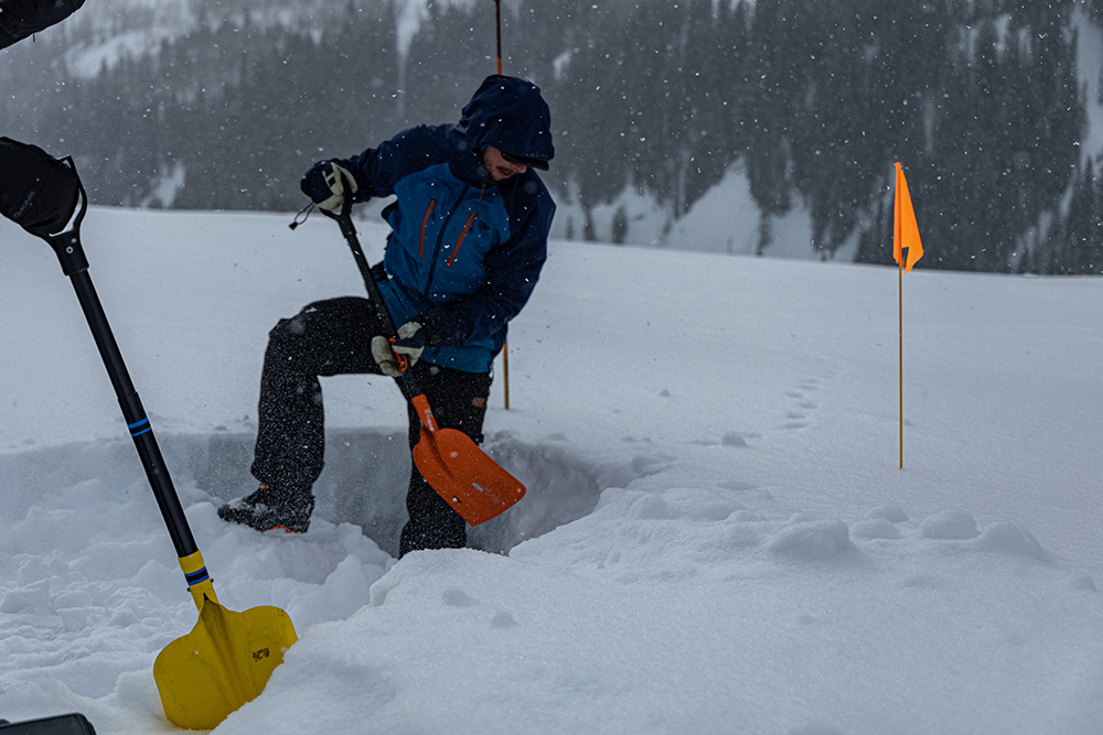 A person stands with one leg in a snow trench and the other leg braced at the edge of the trench. They are digging out the trench with a shovel.