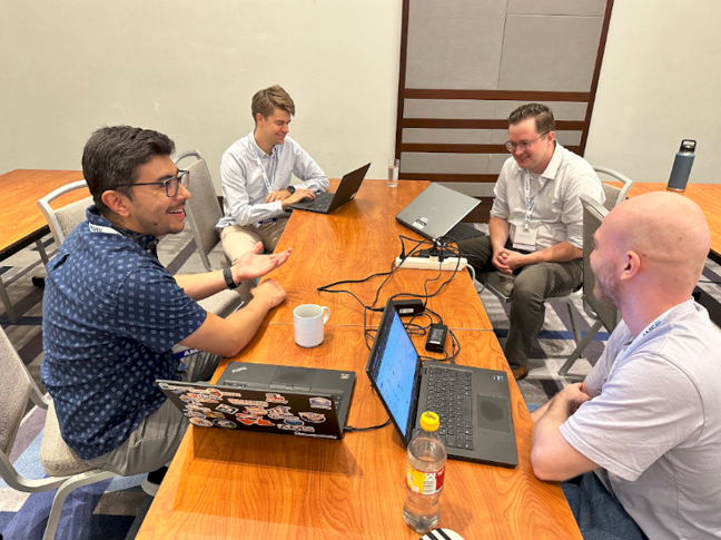 A group of four smiling people sit around a table with open laptops.