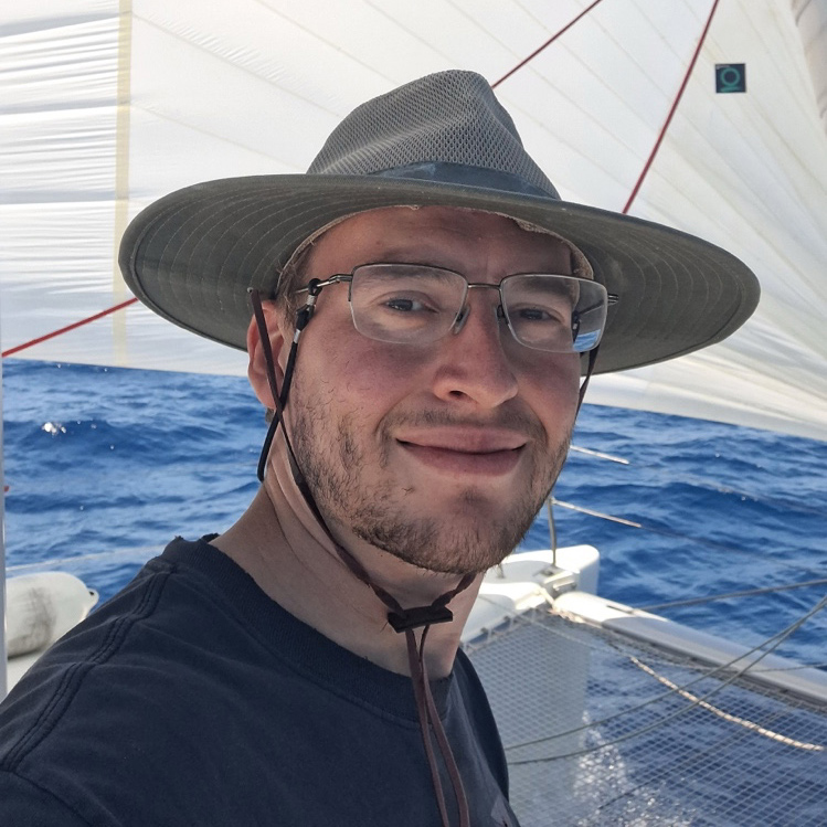 Steven Sharpe wears a sun hat, shirt, and glasses on a sailboat with water behind him.