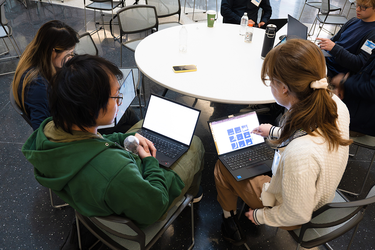 People sit together with laptops open while working around a circular table. One of the people is looking at a neighbor's laptop monitor displaying a square grid of images.