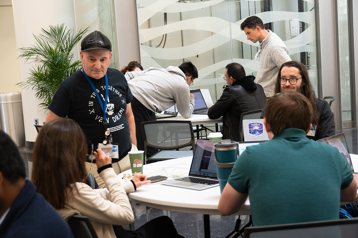 People talk around a circular work table. One of the people is standing up while speaking. The other three people are sitting around the table with laptops open and cups or water bottles next to them.