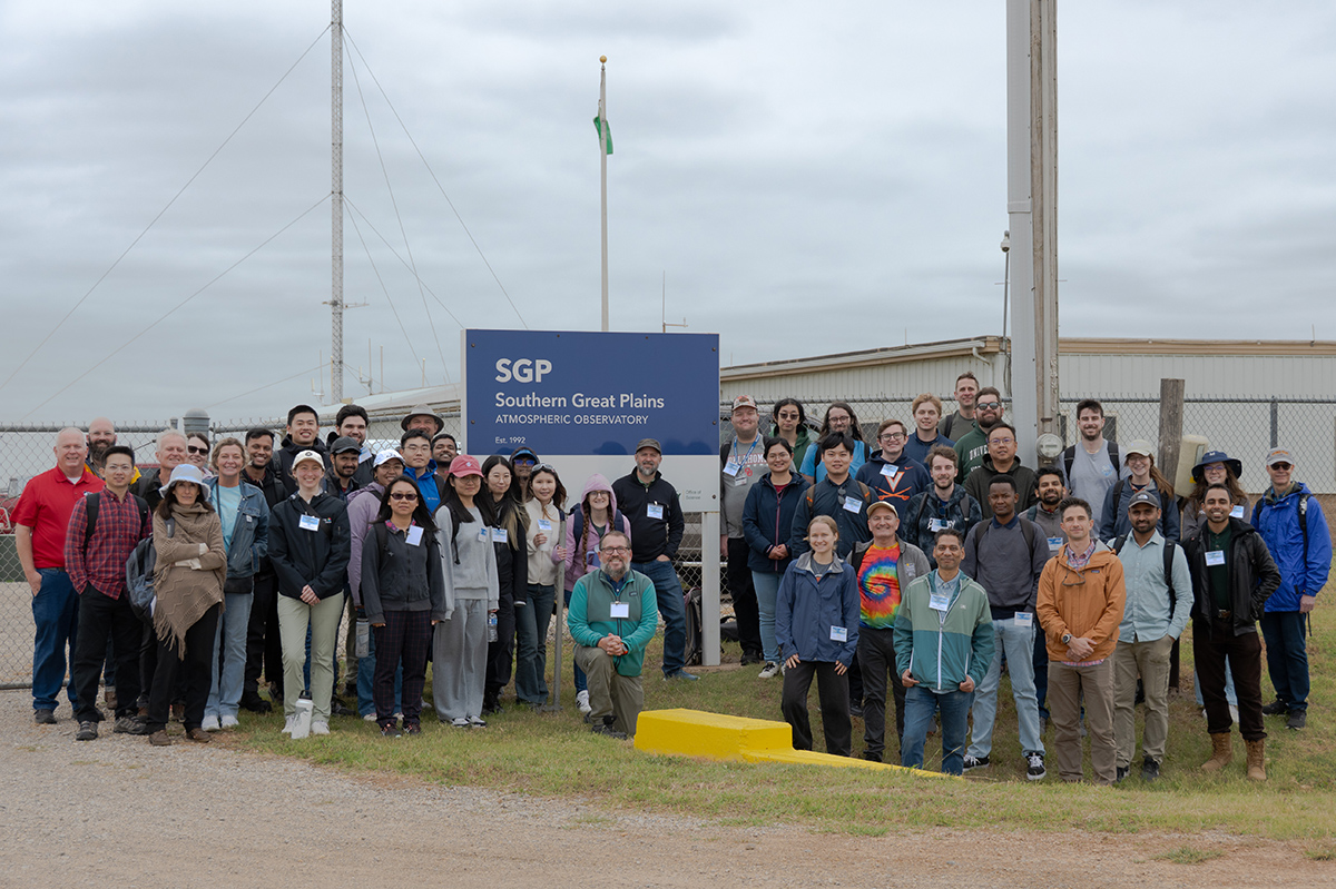 On a cloudy day, a group of people stands around a large sign near a gravel path. The sign says, "SGP, Southern Great Plains atmospheric observatory, Est. 1992." A person at the front of the group is kneeling.