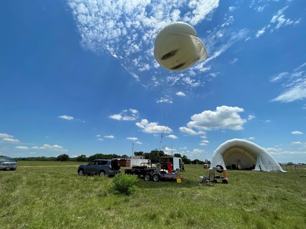 A large balloon is tethered to an instrument trailer with an inflatable shelter behind it and a blue sky with clouds above