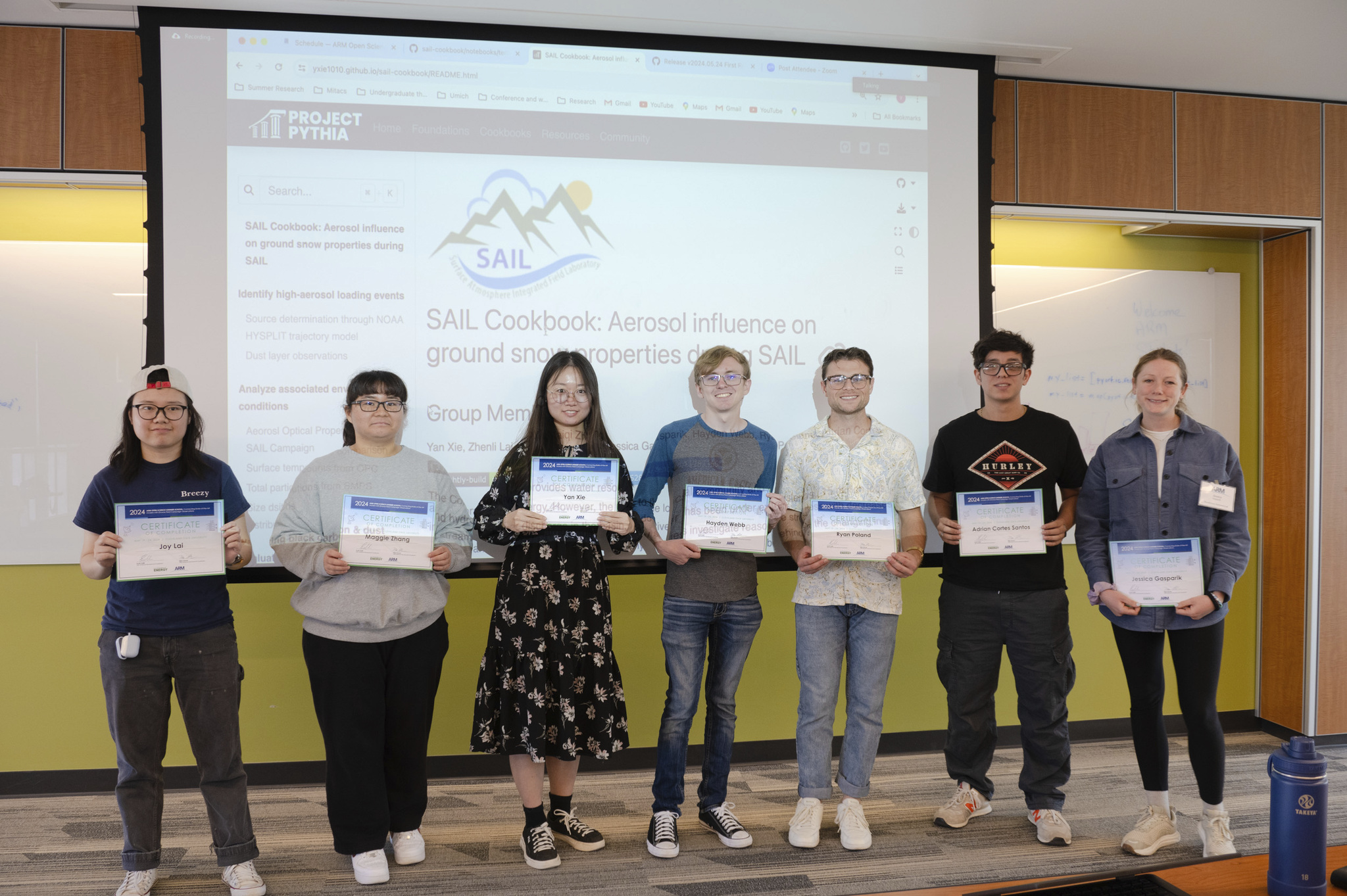 Seven people stand side by side in a room as they hold certificates with their names on them. Behind them is a giant screen that says, "SAIL Cookbook: Aerosol influence on ground snow properties during SAIL."