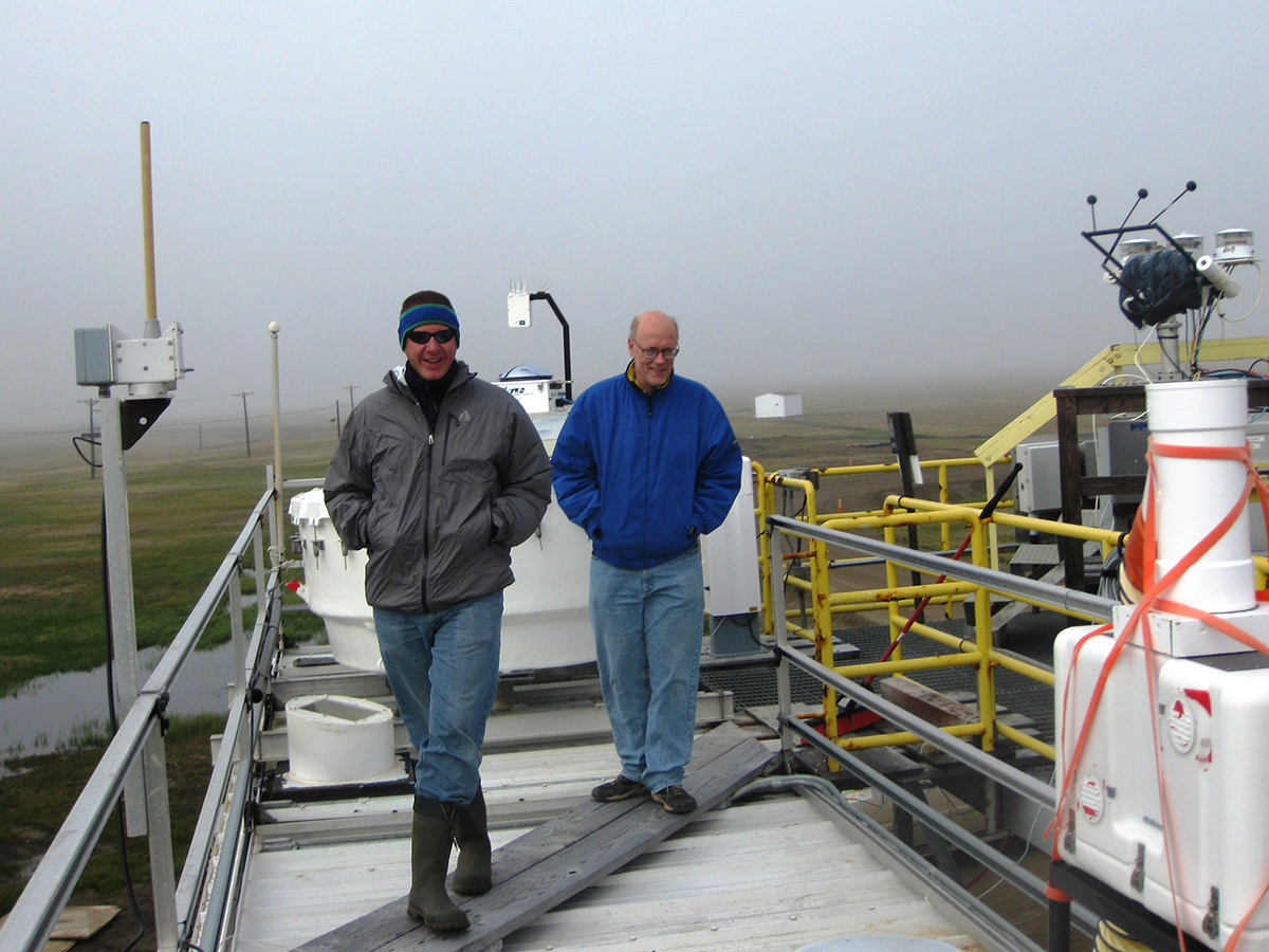 Two people stand on an instrument platform on a foggy day with tundra in the distance.