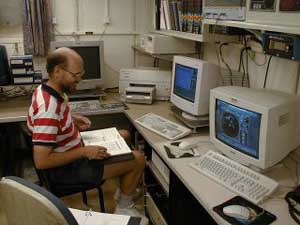 A person looks down at a notebook while sitting in front of two computer terminals.