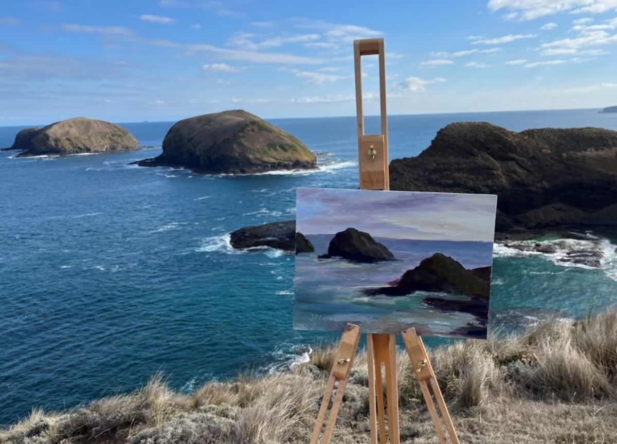 A canvas sits on an easel near a cliff edge. The canvas displays a painted scene that captures the rocks, clouds, and ocean in front of it.