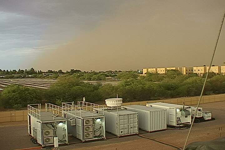 A large dust cloud is seen in the distance beyond atmospheric instrumentation and a modern academic building. A line of trees separates the instruments and the building.
