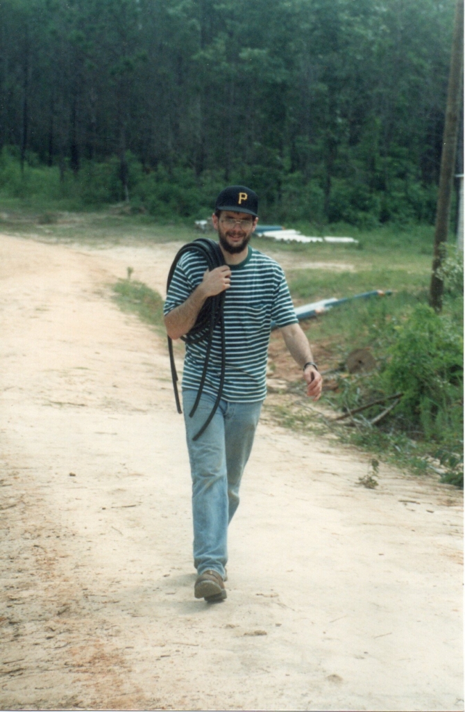 Jim Mather is dressed in a Pittsburgh Pirates cap, striped shirt, blue jeans, and boots holding tubing on his right shoulder. He is walking on a smooth dirt path with forest behind him and different vegetation along the side of the path.