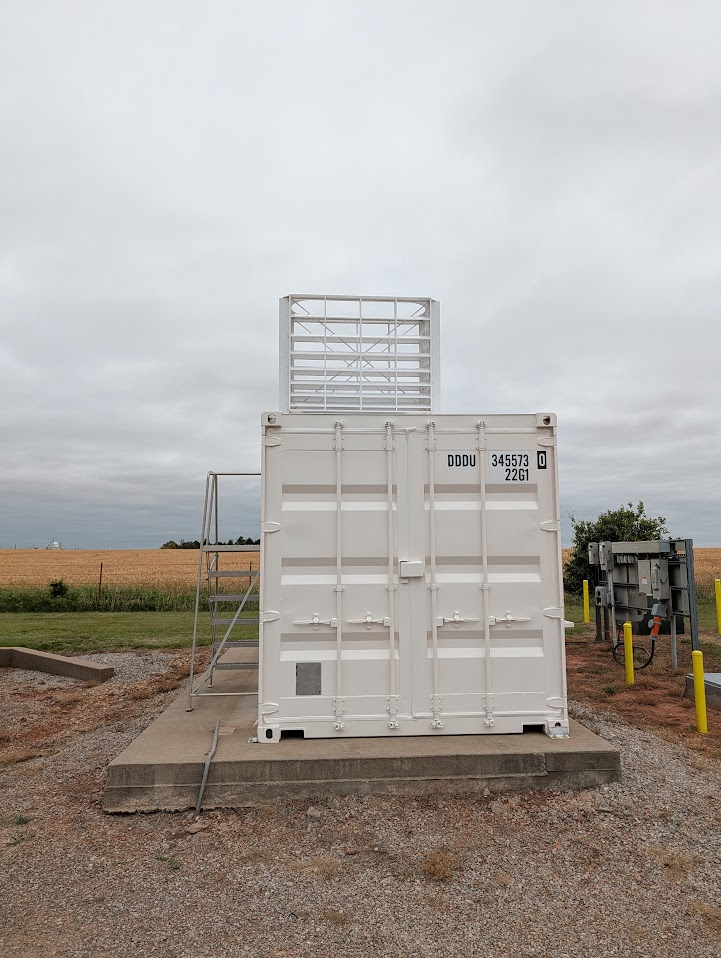 A sunshade for ARM's high-spectral-resolution lidar protrudes from the top of an instrument container. Cropland is seen in the distance on a cloudy day.