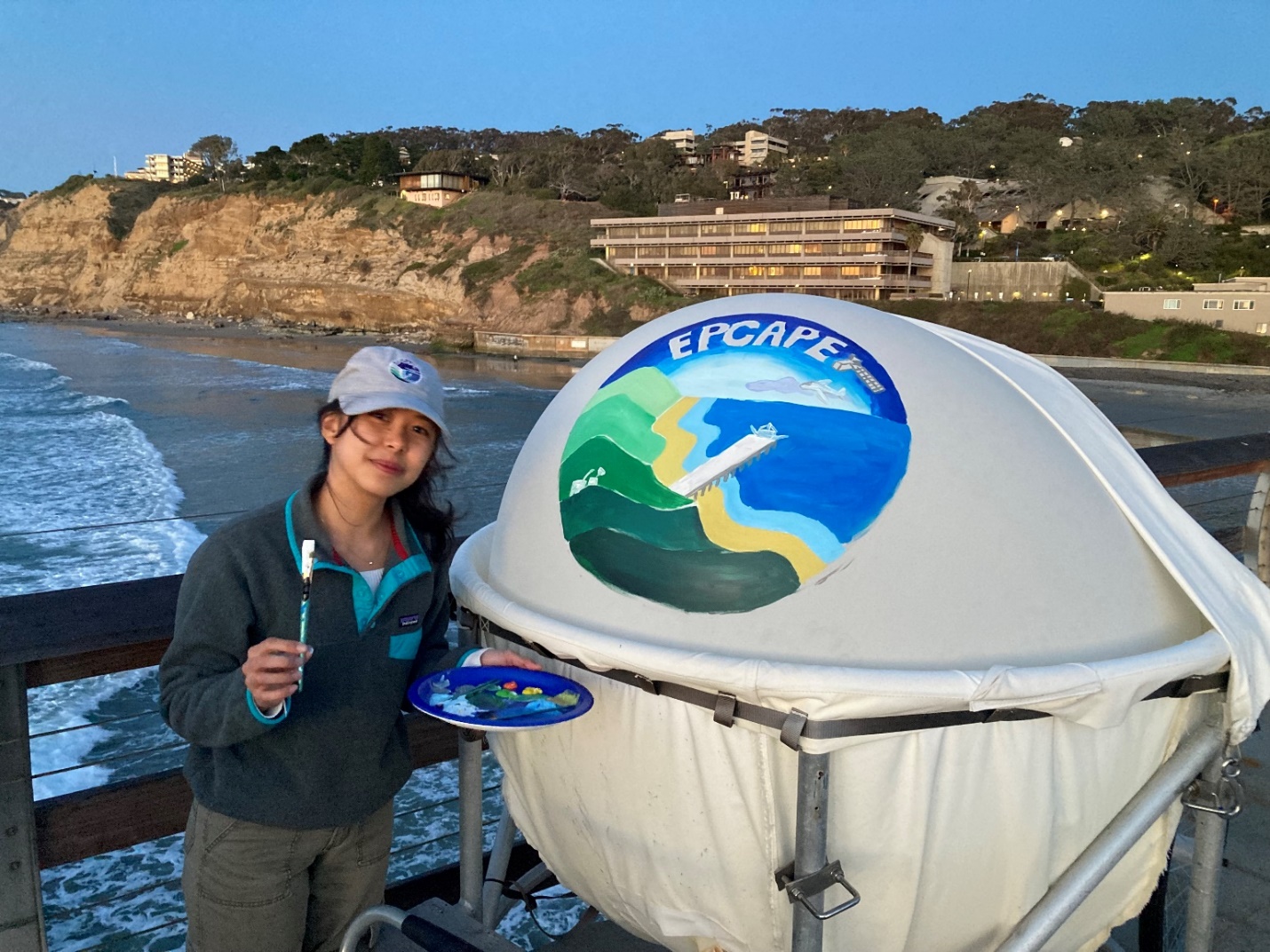 A person wearing a cap holds a paintbrush in one hand and a palette with different paint colors in the other. They are standing next to a weather balloon painted with a scene from EPCAPE. Below the person on the pier, ocean waves crash into the shoreline.