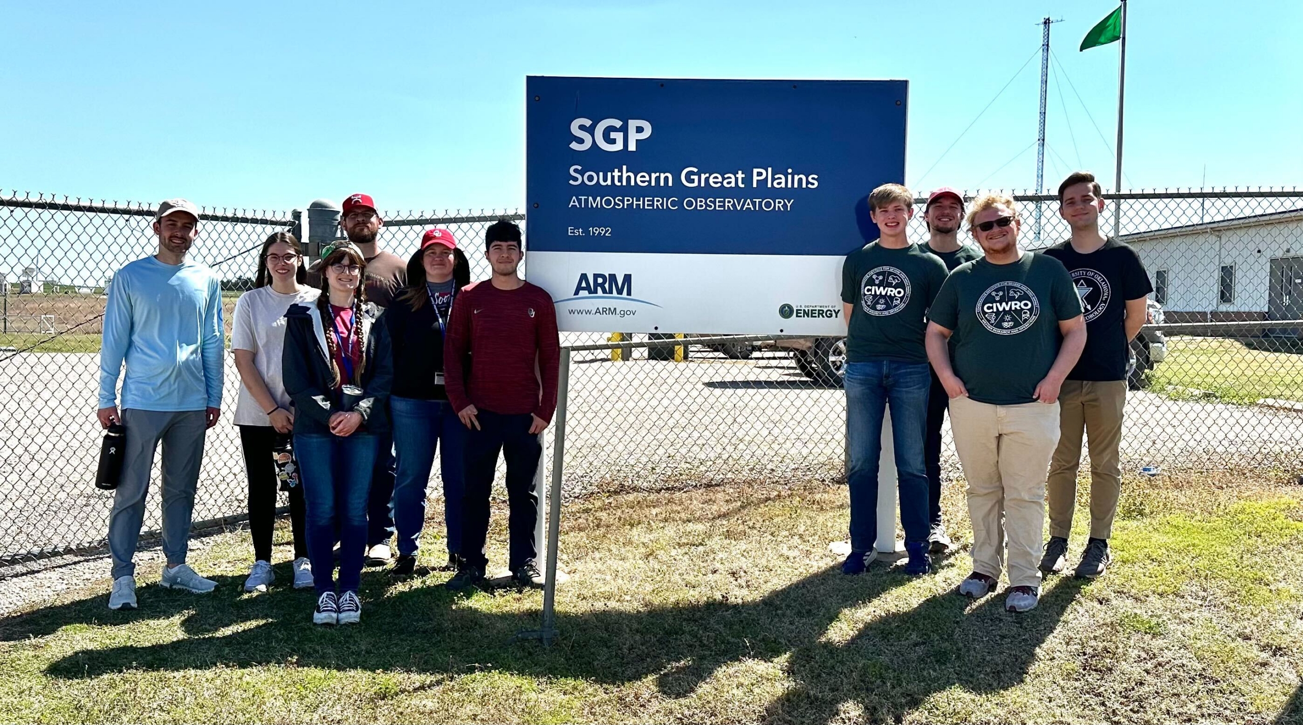 Six people stand on one side of a entrance sign for ARM's Southern Great Plains atmospheric observatory, and four people stand on the other side.