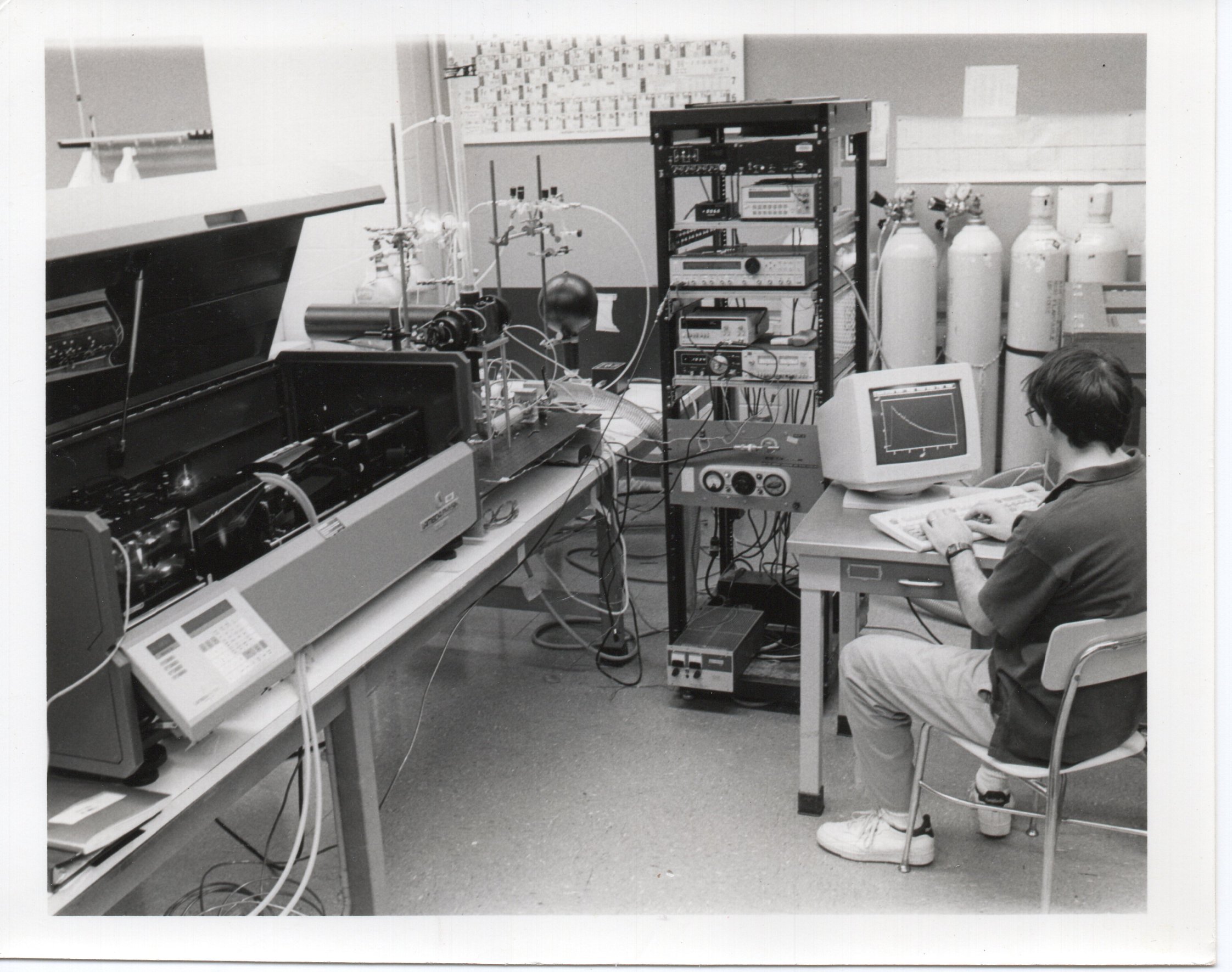 In a scientific laboratory, Jim Mather types on a keyboard looking at a graph on a computer monitor. The lab has different kinds of systems and wiring throughout.