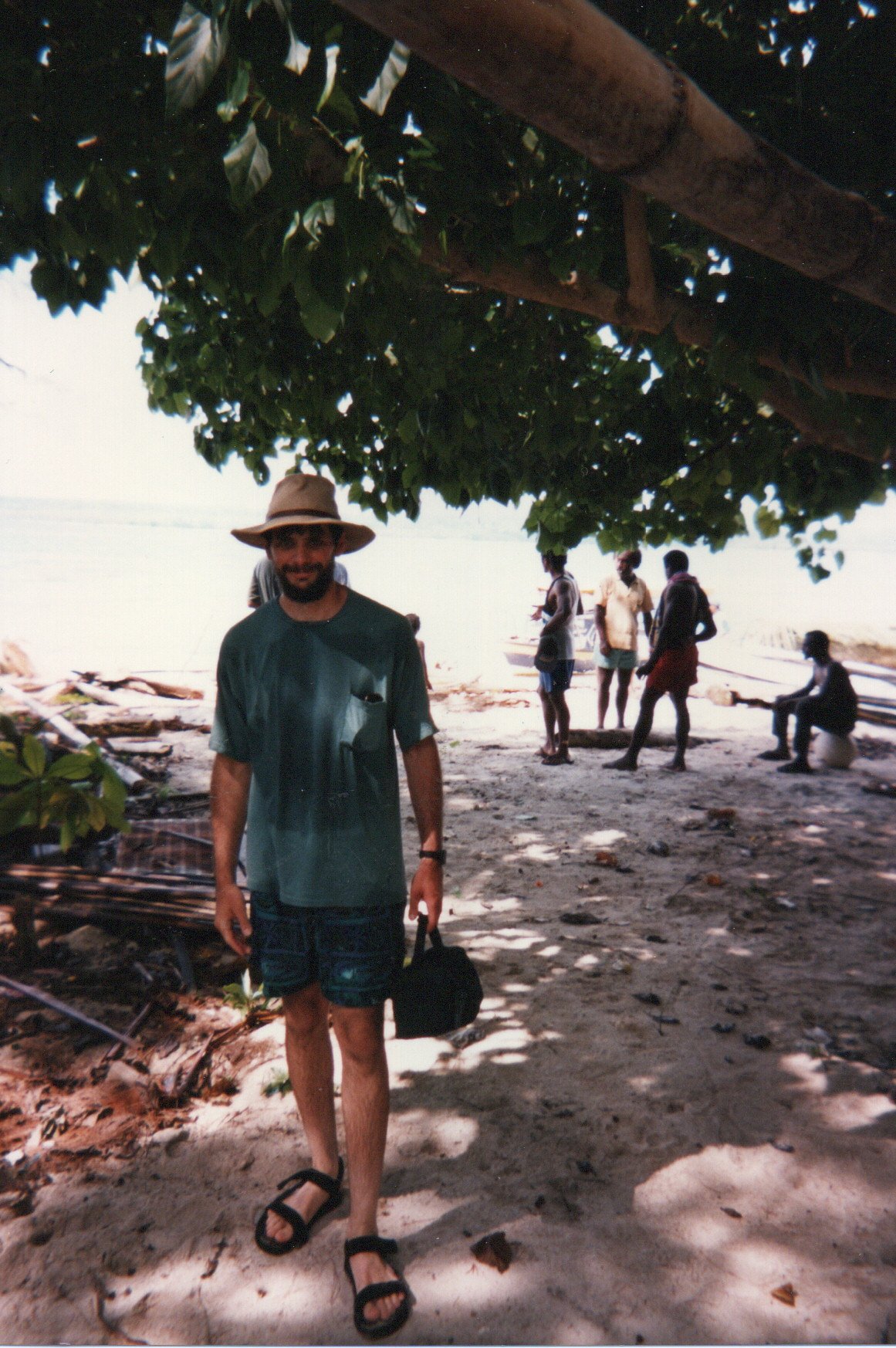 Jim Mather is dressed in a tan hat, green T-shirt, shorts, and sandals underneath a tree. He is holding a small bag by the handle. Others behind him are wearing different kinds of beach apparel.